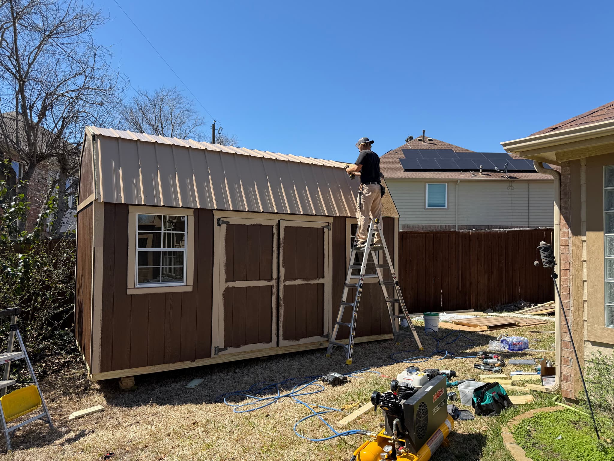 A builder on a ladder installing the metal roof on a finished-looking on-site shed.