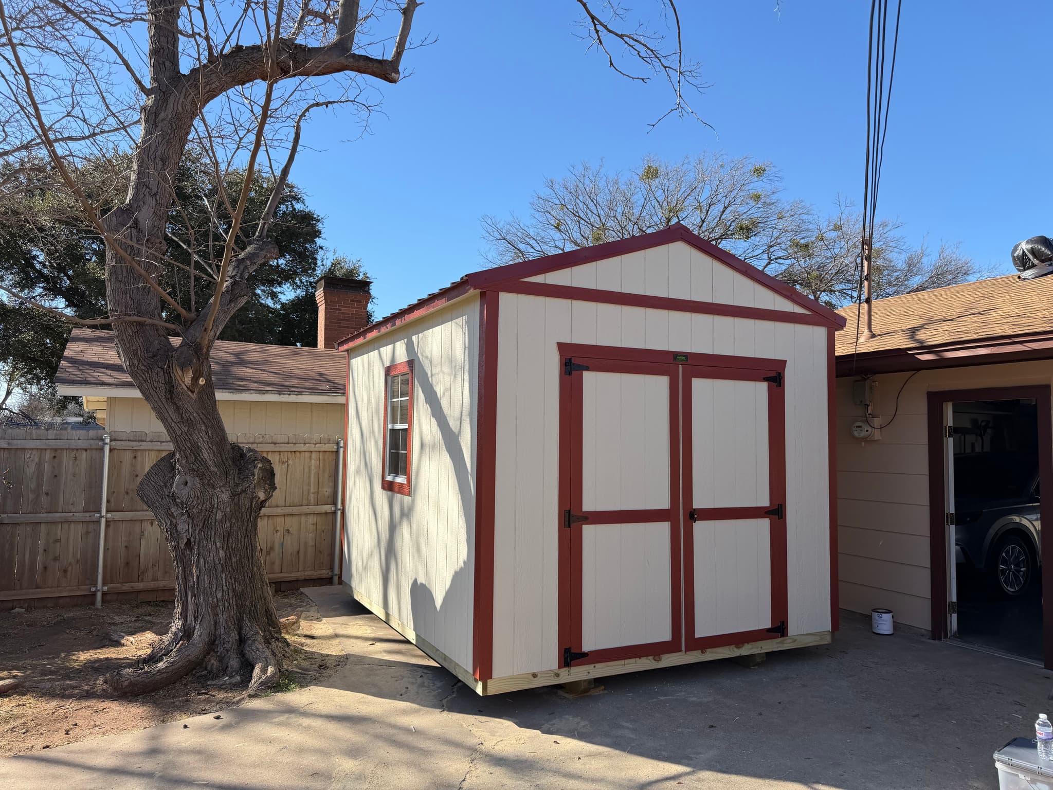 Finished cream 8x12 utility shed with red metal roof and red trim in a tight backyard.